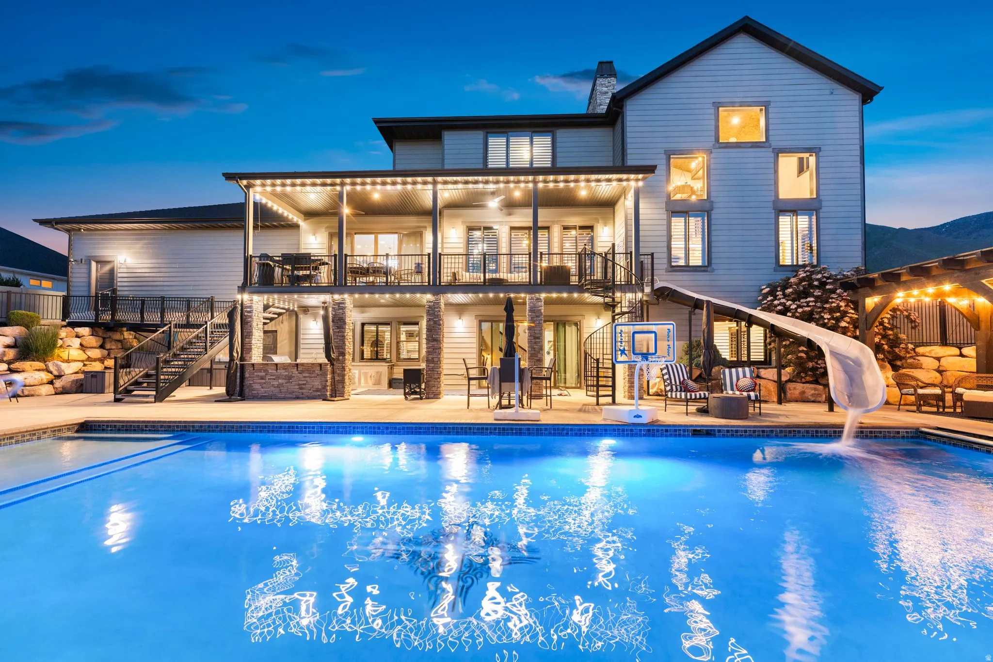 Extensive backyard featuring a swimming pool with a built-in slide, a covered patio with string lights, a second-story balcony, a spiral staircase, and stone-clad architectural elements
