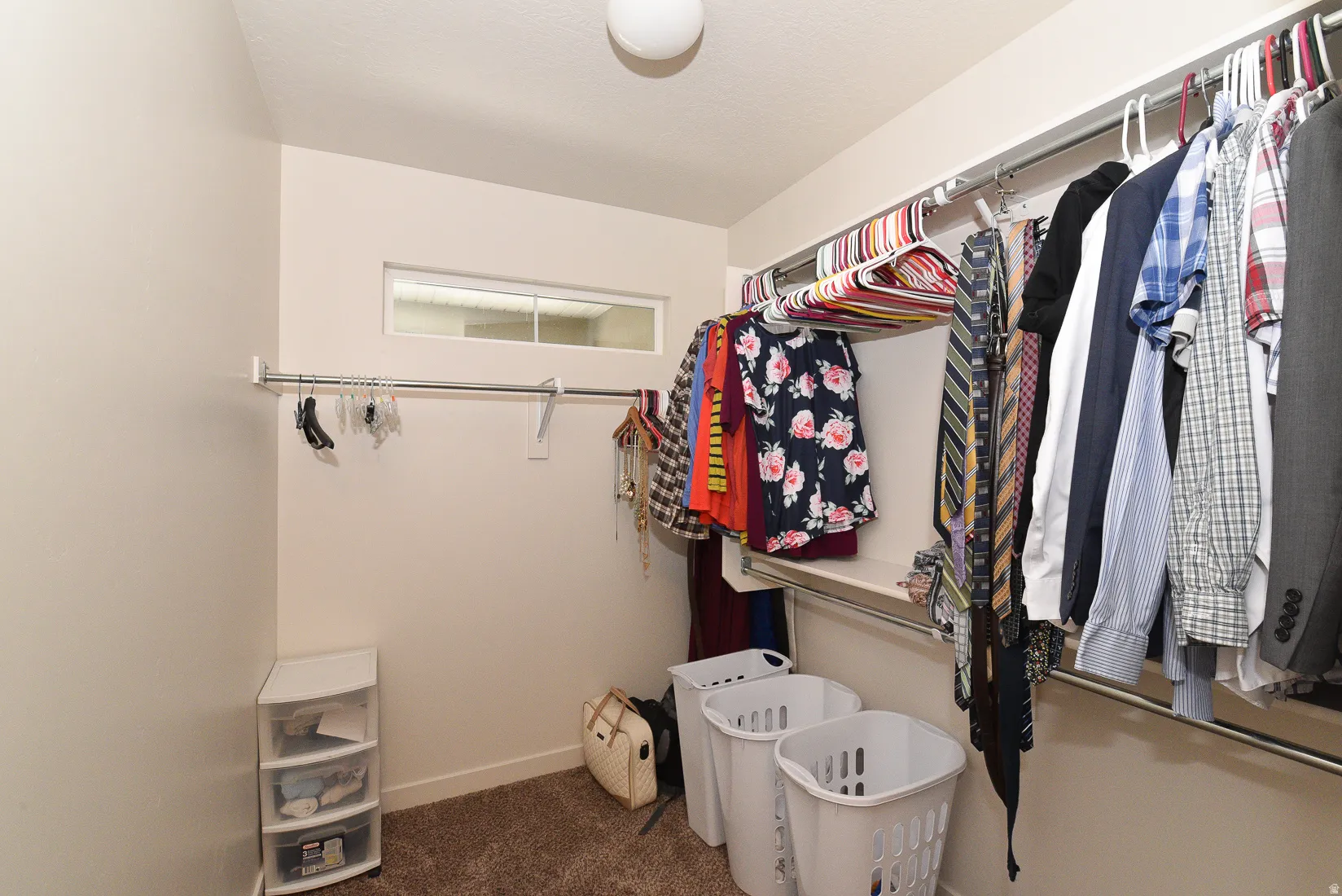 Walk-in closet with carpet flooring, multiple hanging rods, and a high-set horizontal window