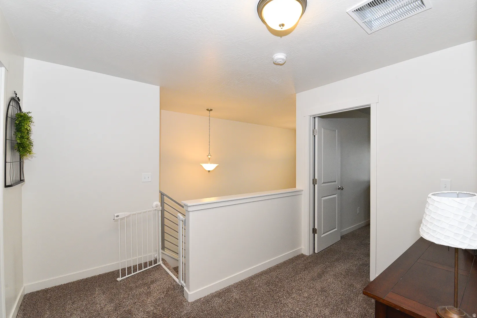 Carpeted upper-level landing featuring a white painted railing with metal spindles, a flush-mount ceiling light, a recessed ceiling vent, and a hallway leading to an interior door