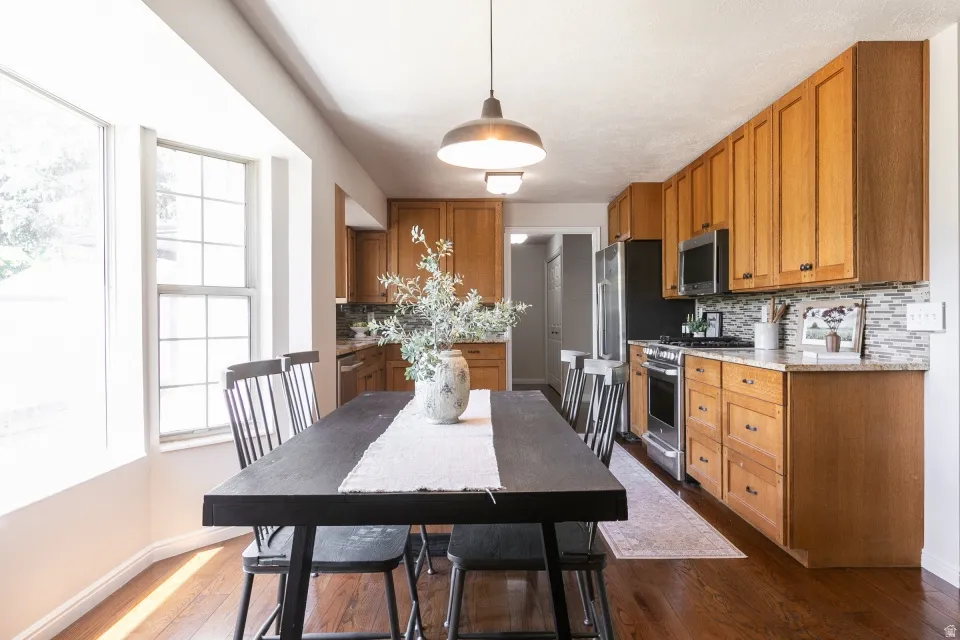 Open-concept kitchen and dining area featuring wood-finish flooring, ample natural light from large windows, wood cabinetry, stone countertops, and a mosaic tile backsplash
