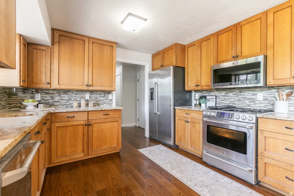 Kitchen featuring wood cabinetry, stainless steel appliances, granite countertops, a tiled backsplash, and wood-finish flooring