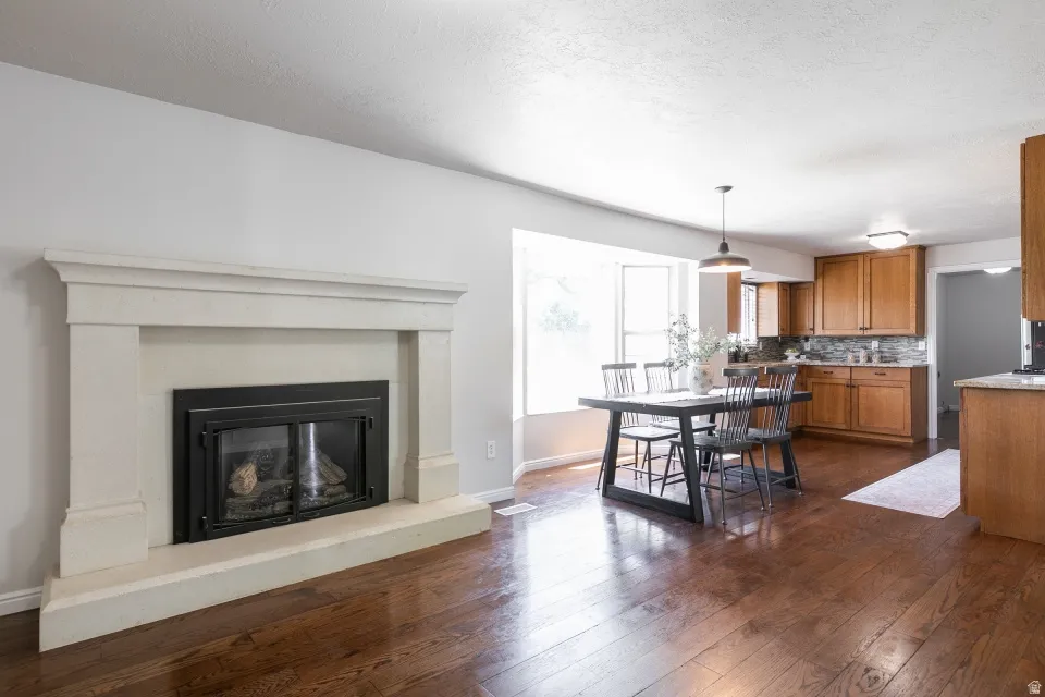 Open-concept living area featuring a stone-look fireplace, wood-finish flooring, a bay window, and kitchen cabinetry with a mosaic tile backsplash