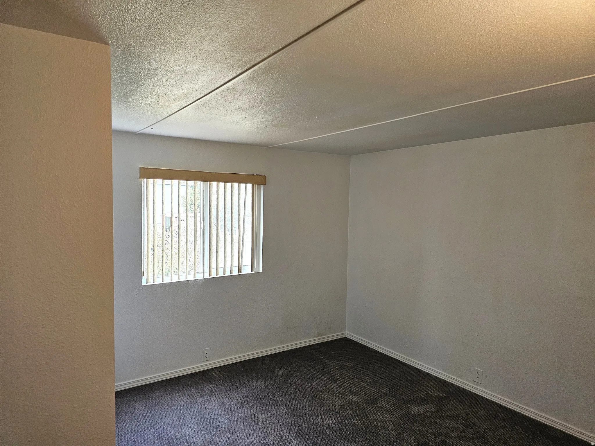 Interior room featuring a window with vertical blinds, light-colored walls, dark carpeting, and white baseboards