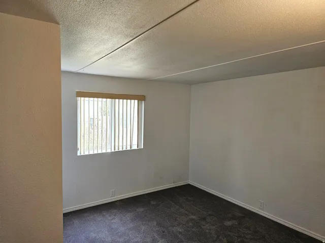 Interior room featuring a window with vertical blinds, light-colored walls, dark carpeting, and white baseboards