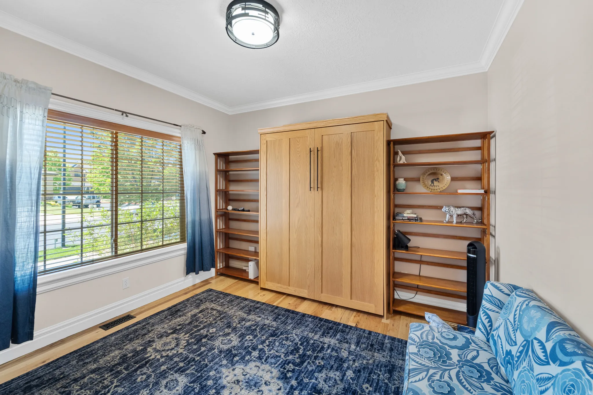 Room featuring light wood-finish flooring, a large window with horizontal blinds, built-in wooden shelving, a built-in wall bed, and crown molding