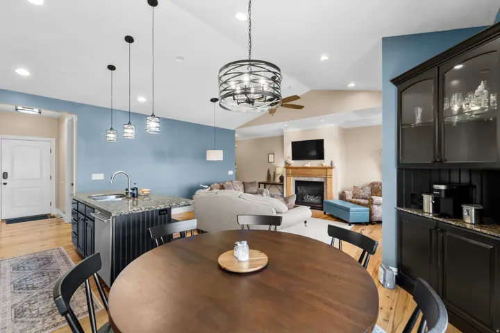 Open-concept living area featuring wood-finish flooring, a kitchen island with stone countertop, a built-in beverage bar with glass-front cabinetry, a fireplace with a wood mantle, and vaulted ceilings