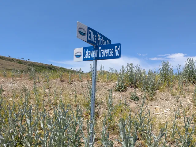 Road sign indicating "Elks Ridge Rd" and "Lakeview Traverse Rd" on a hillside with sparse vegetation under a clear blue sky