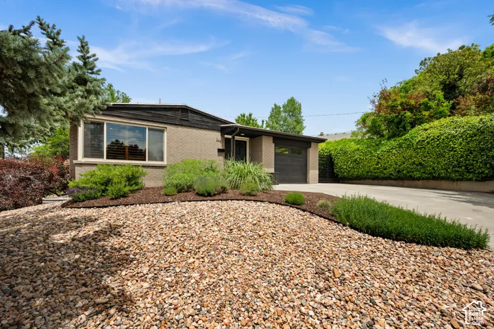 Brick exterior featuring a dark garage door, large front window with plantation shutters, low-maintenance gravel landscaping, and mature shrubbery