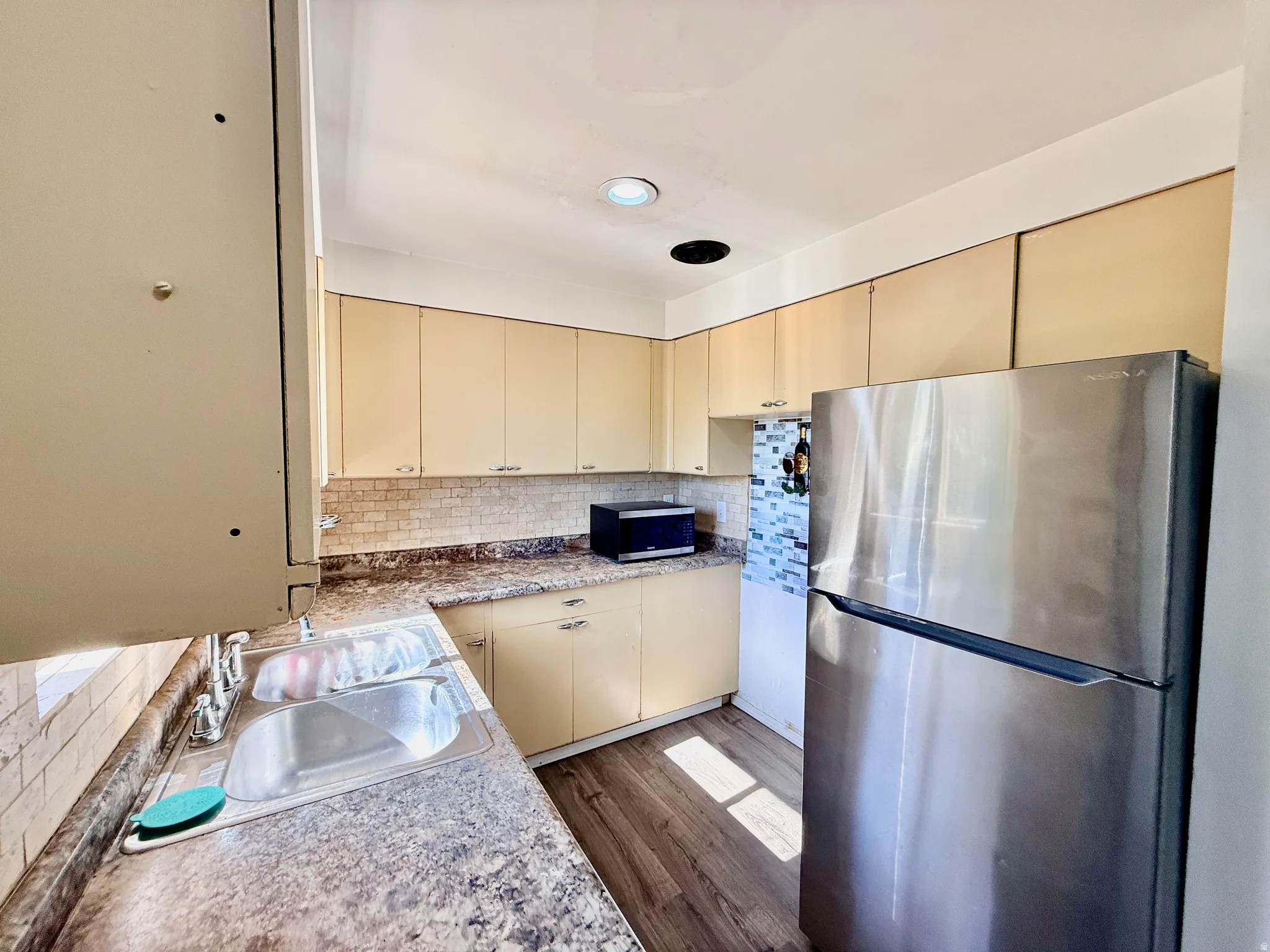 Kitchen featuring a double stainless steel sink with a chrome faucet, light wood-finish cabinetry, patterned backsplash, and dark wood-finish flooring