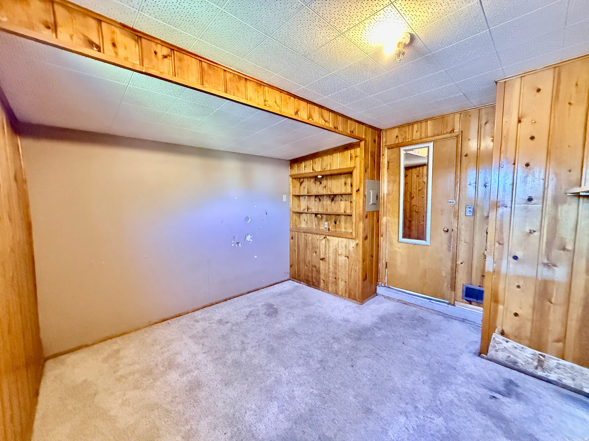 Carpeted room featuring wood paneling, a built-in shelving unit, a door with an integrated mirror, and a drop ceiling