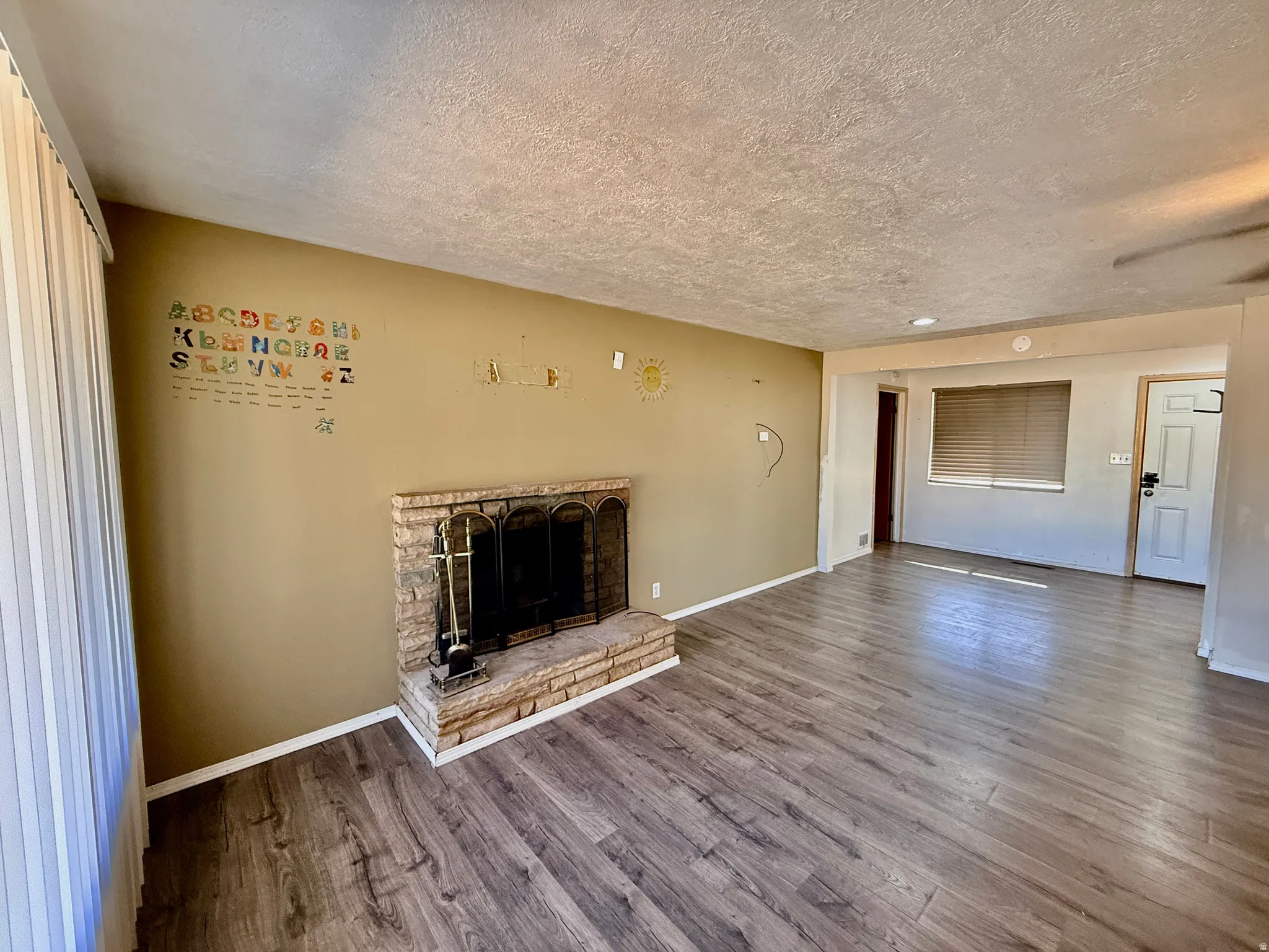 Living area featuring wood-finish flooring, a brick fireplace with a black screen, a ceiling fan with integrated lighting, and horizontal blinds on the window