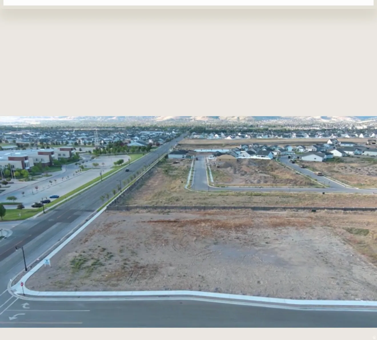 Undeveloped land parcel with paved road frontage, established residential areas in the background, and distant mountain ranges
