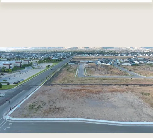 Undeveloped land parcel with paved road frontage, established residential areas in the background, and distant mountain ranges