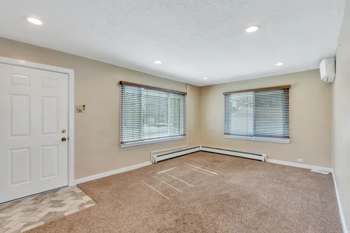 Living area featuring recessed lighting, a white panel door, two windows with blinds, baseboard heating, and a wall-mounted unit