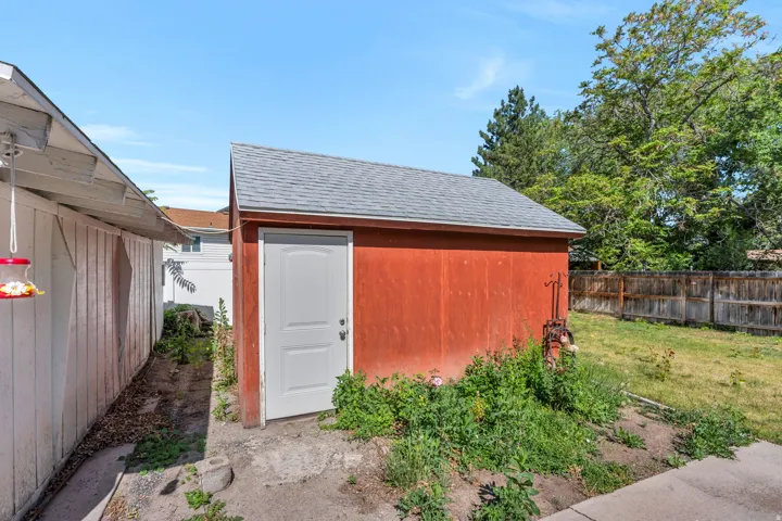 Detached red storage shed with a white entry door and a gray shingled roof, featuring a surrounding grassy area and a wood plank fence