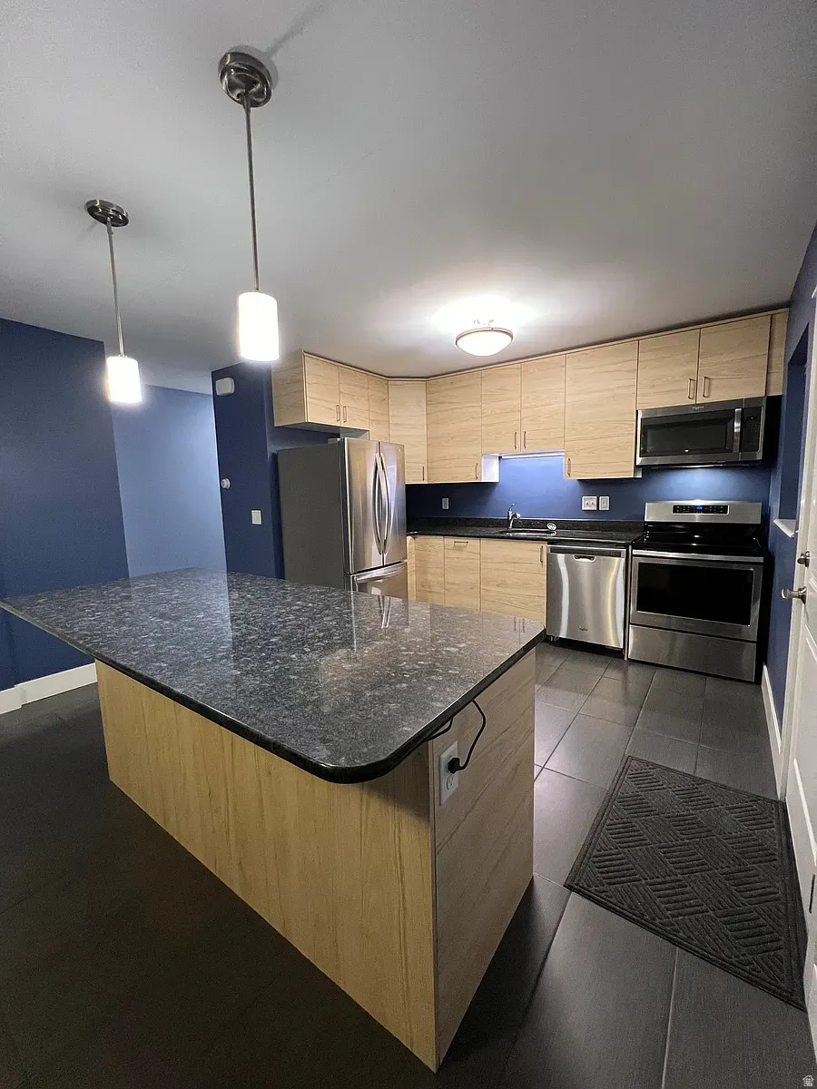 Kitchen with light wood finish cabinets, stainless steel appliances, a kitchen island, and dark stone counters