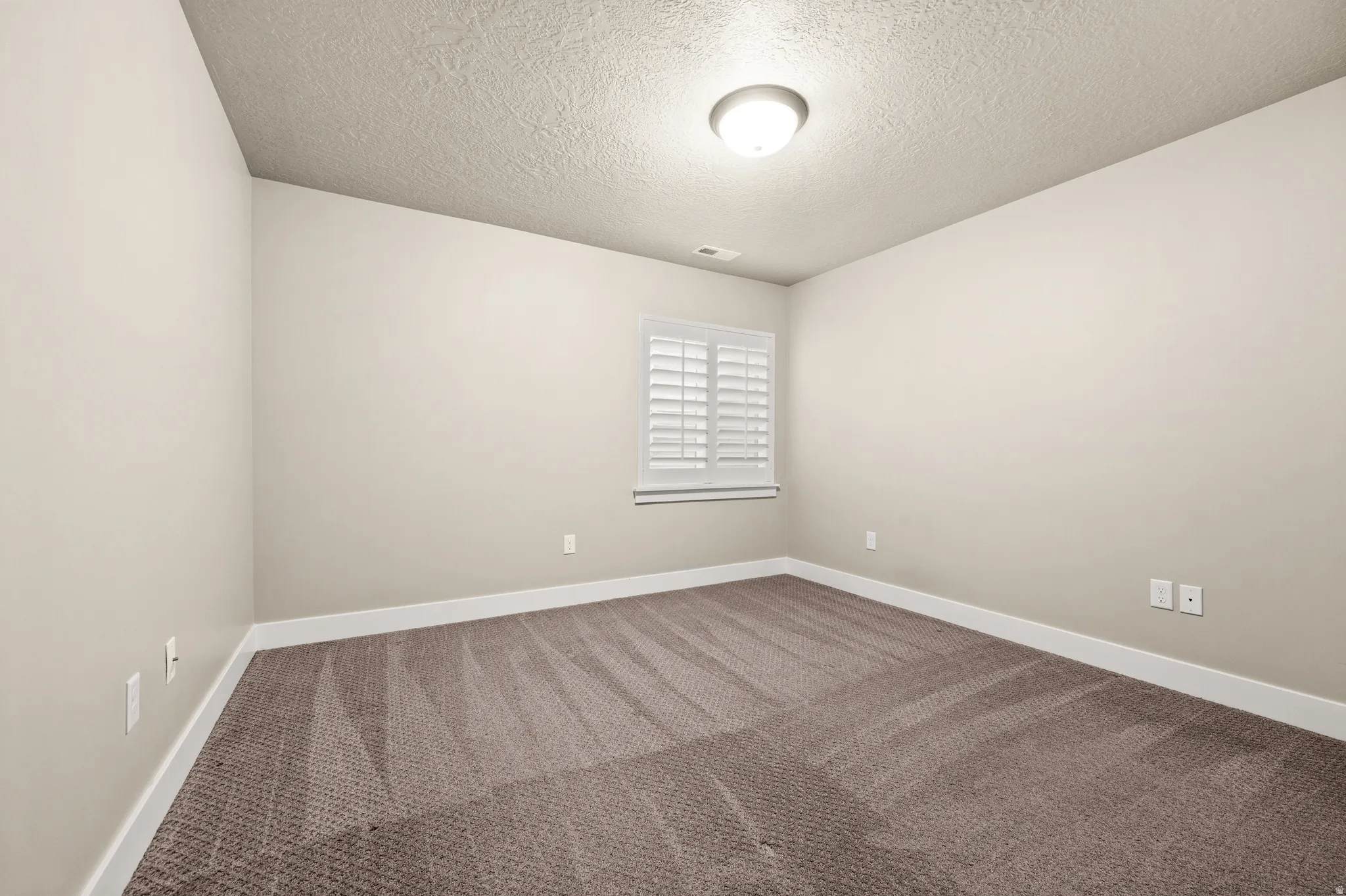 Neutral-toned room featuring a window with white plantation shutters, textured ceiling, and a flush mount ceiling light