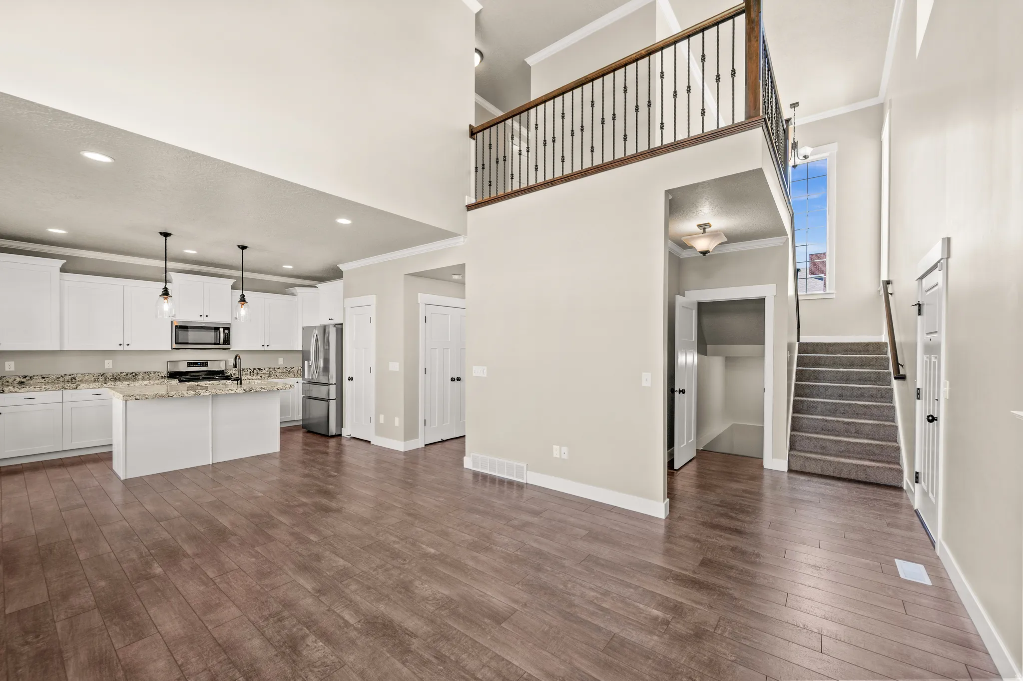 Open-concept living space with wood-finish flooring, a kitchen featuring white cabinetry and stainless steel appliances, a multi-level ceiling, and a staircase with a wrought iron railing
