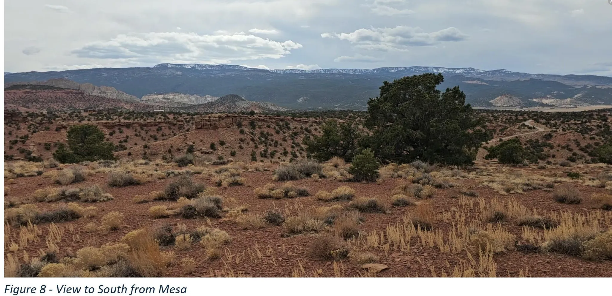 Expansive landscape featuring red soil, desert vegetation, distant mountains, and varied topography under a cloudy sky