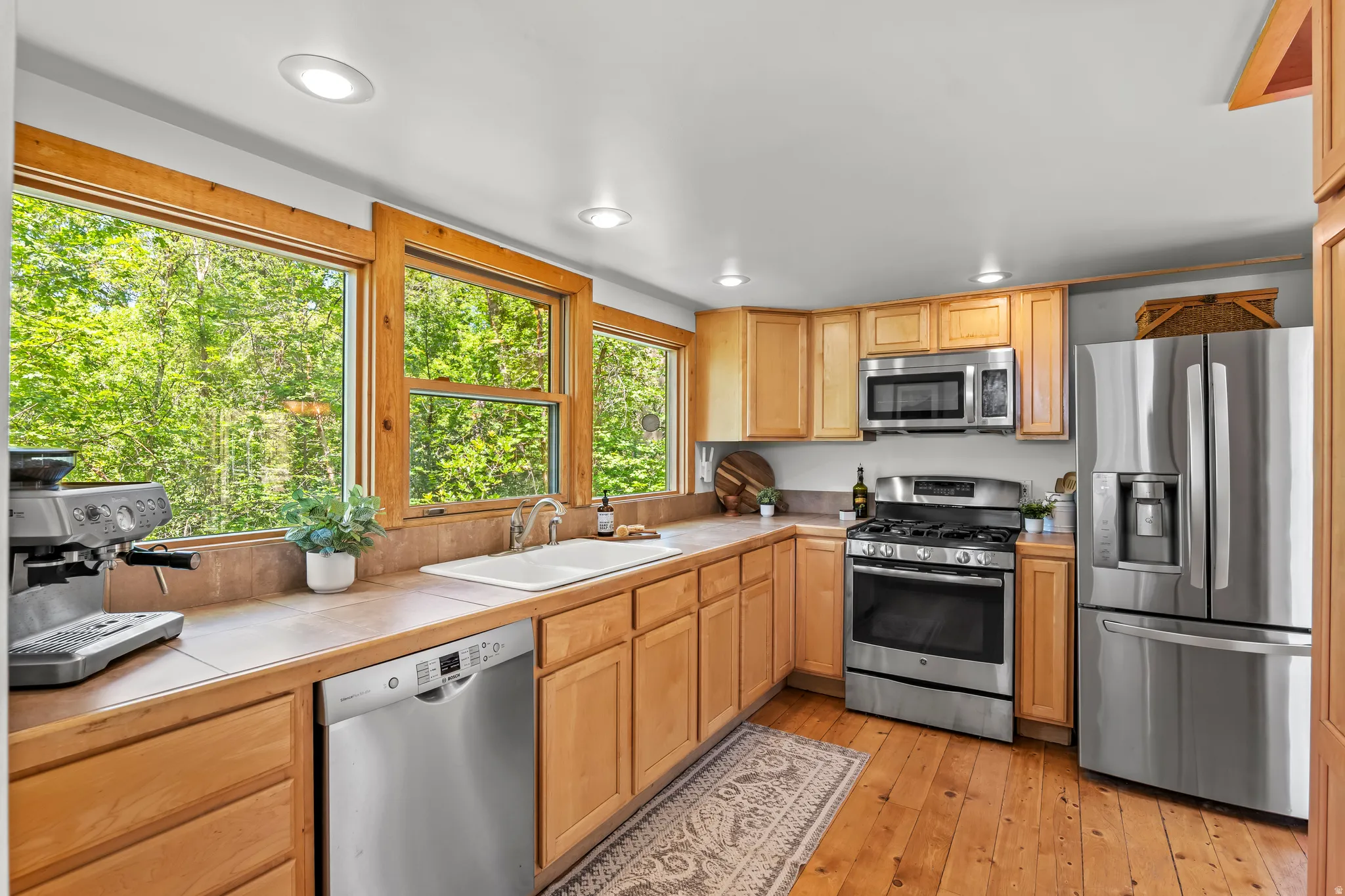 Kitchen featuring abundant natural light from large windows, wood-finish flooring, stainless steel appliances, light wood cabinetry, and recessed lighting