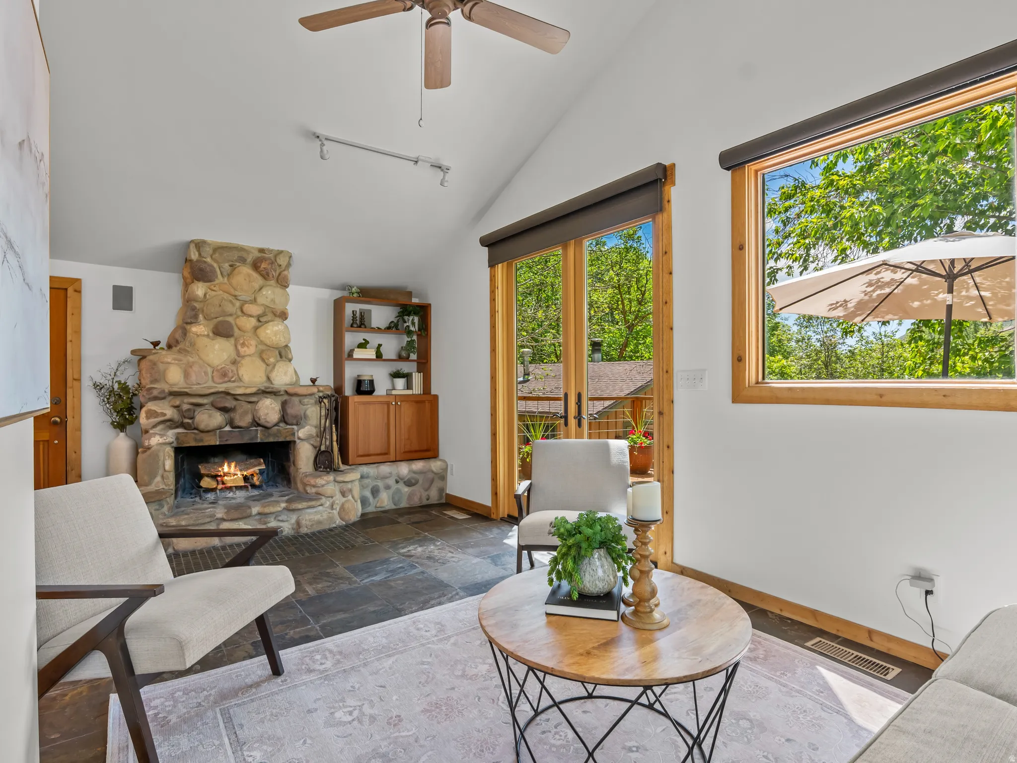 Vaulted ceiling with wood-finish ceiling fan, stone fireplace, slate flooring, and expansive windows with wood trim