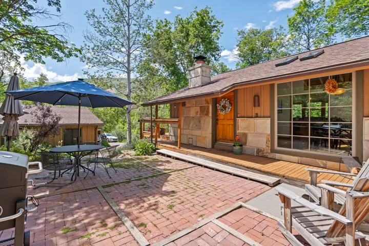 Brick paver patio with wood deck accents, featuring a large picture window, stone and wood exterior siding, a natural wood entry door, and a prominent stone chimney