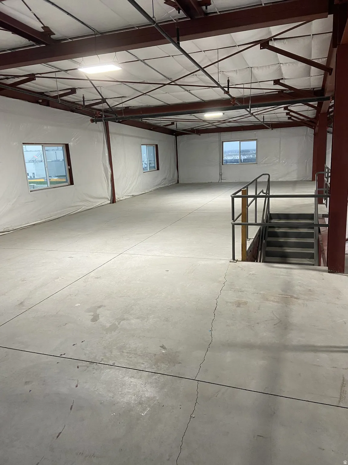 Spacious open-concept commercial interior featuring exposed structural beams, concrete flooring, multiple windows, and fluorescent lighting