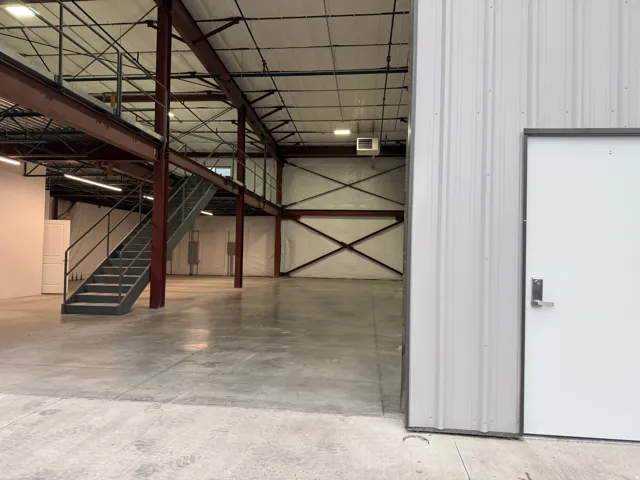 Spacious industrial interior featuring a concrete floor, exposed structural beams, a mezzanine level with metal stairs, overhead lighting, and insulated walls