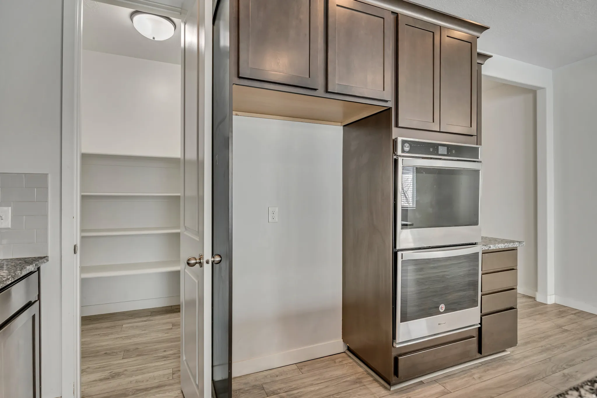 Kitchen featuring wood-finish flooring, built-in double ovens, dark wood cabinetry with crown molding, granite-style countertops, and a full-height pantry with multiple shelves