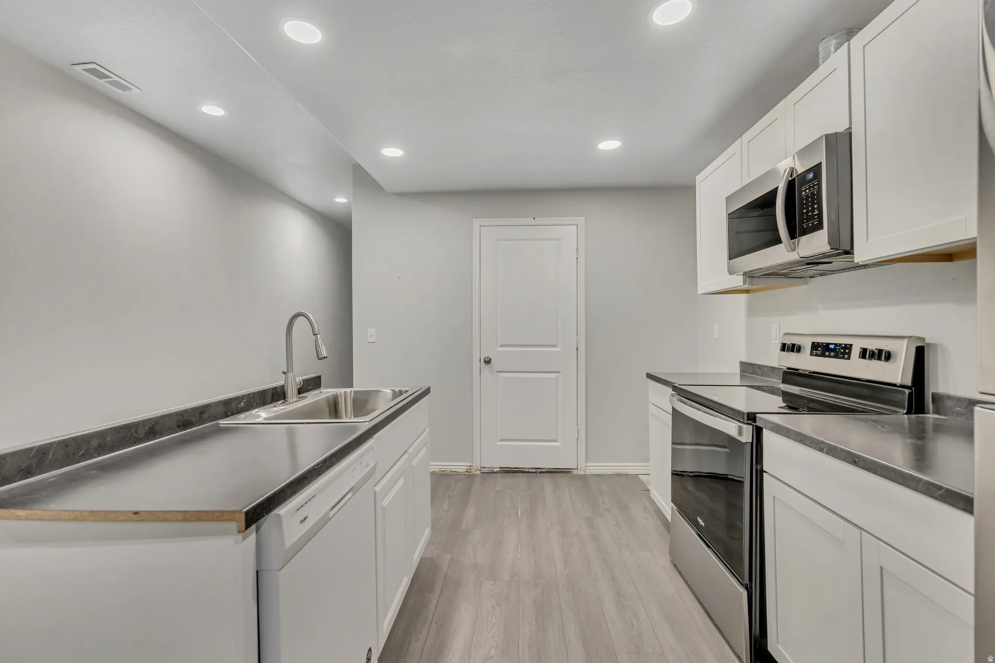 Modern kitchen featuring white cabinetry, dark countertops, stainless steel appliances including a microwave and range, a stainless steel sink with a gooseneck faucet, and wood-finish flooring