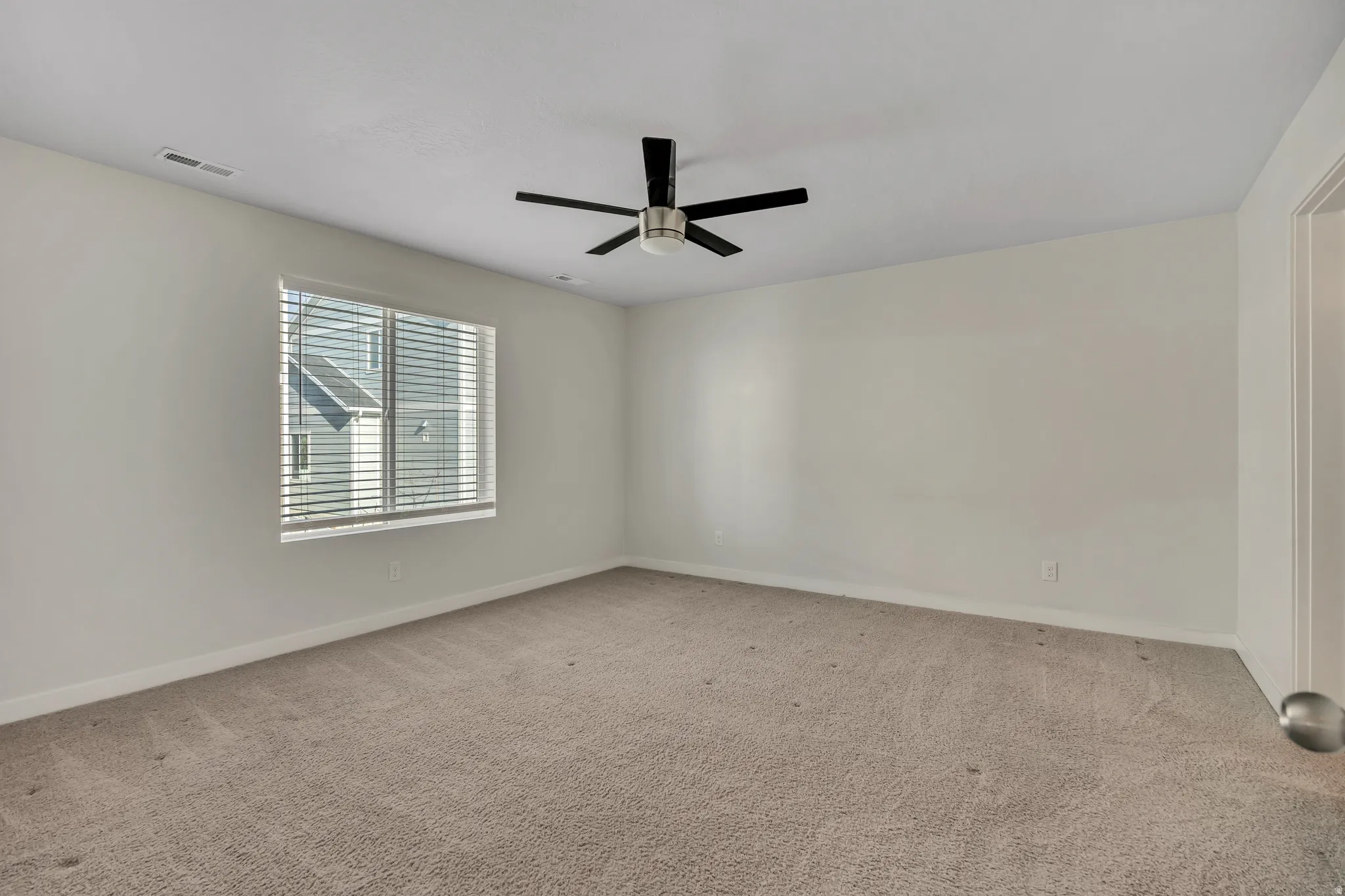 Spacious room featuring neutral carpeting, light-toned walls, and a ceiling fan with black blades