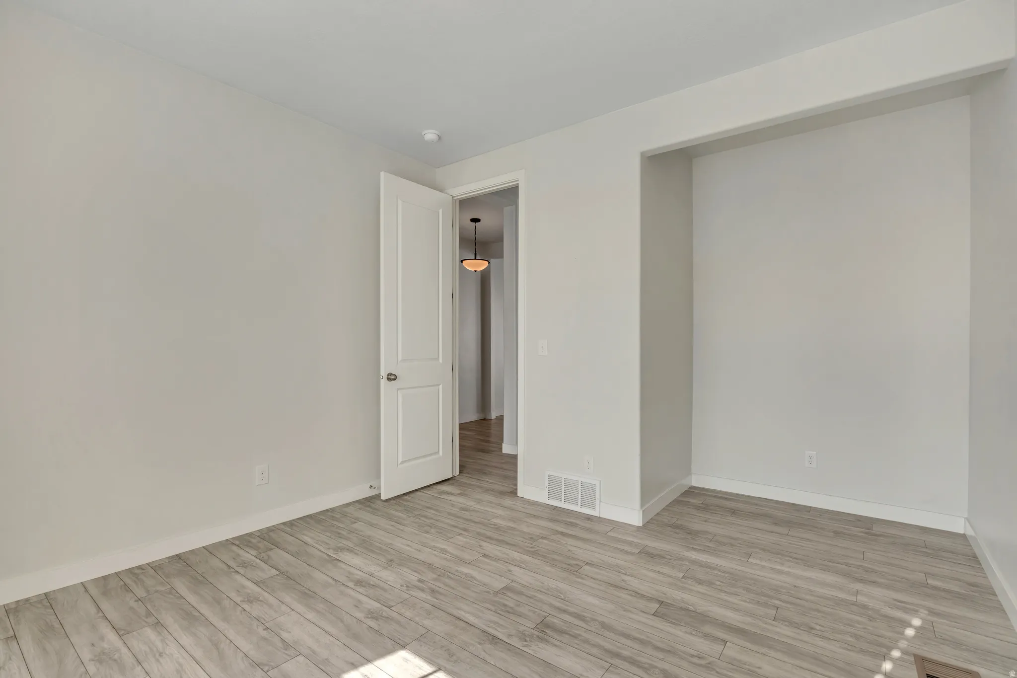 Spacious interior room featuring light wood-finish flooring, white baseboards, and a white interior door with a silver-tone knob