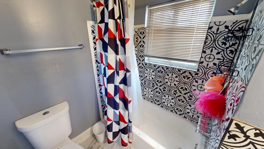 Bathroom featuring a white porcelain toilet, a shower with patterned wall tile, and a window with horizontal blinds