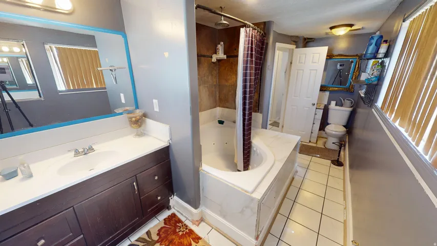 Bathroom featuring a built-in jetted tub with tiled surround, a vanity with a white countertop and dark wood-finish cabinetry, a wall-mounted mirror, and white tile flooring