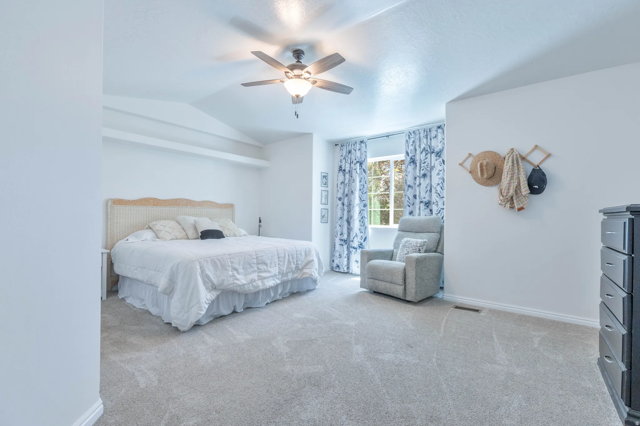 Spacious carpeted room featuring a vaulted ceiling with a ceiling fan, a large window with drapes, and white baseboards