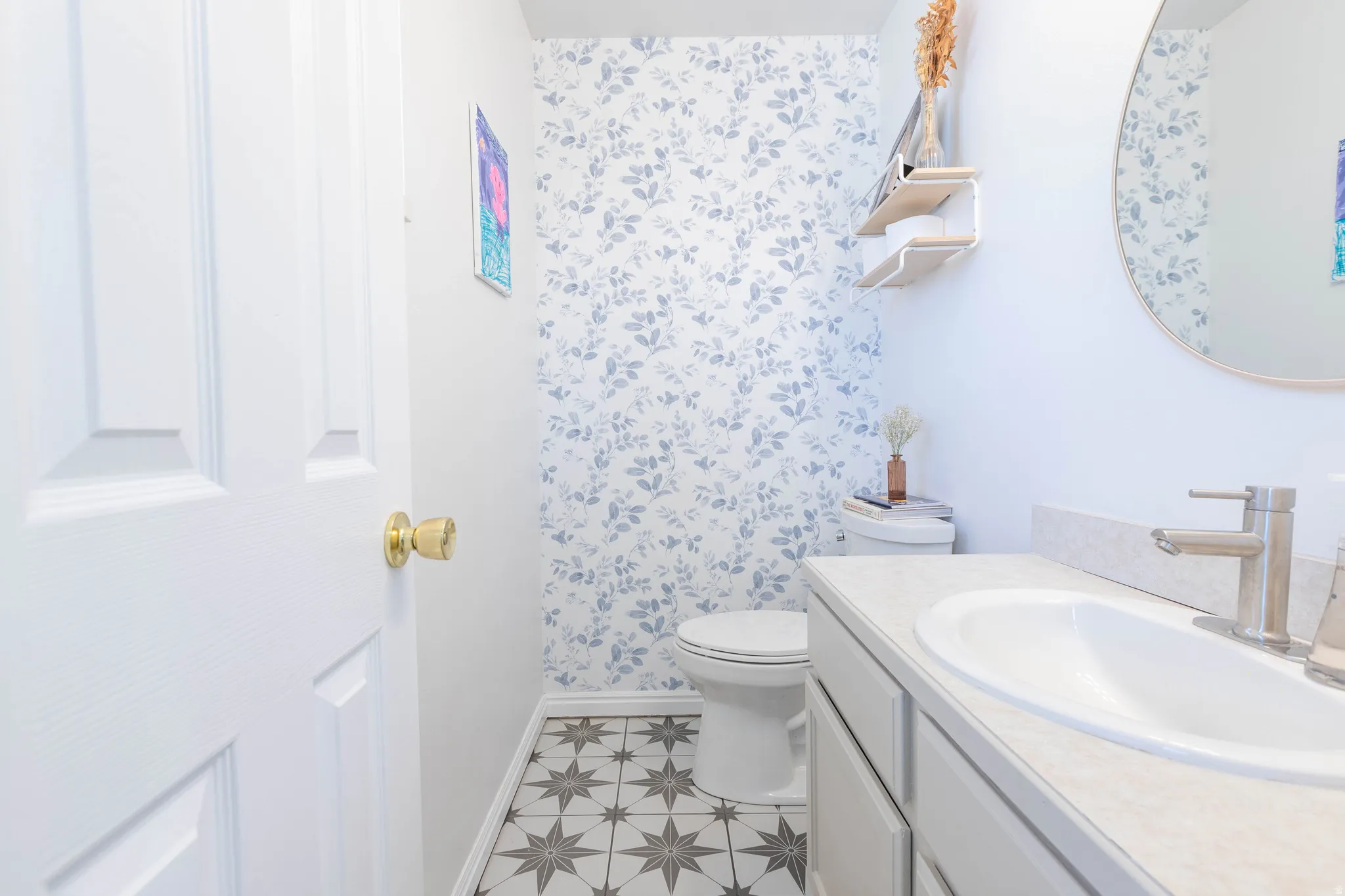 Powder room featuring patterned floor tile, a single vanity with a white countertop and integrated sink, a large round mirror, and a wallpaper accent wall
