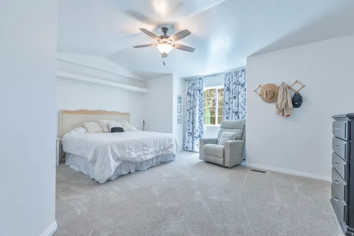 Spacious carpeted room featuring a vaulted ceiling with a ceiling fan, a large window with drapes, and white baseboards