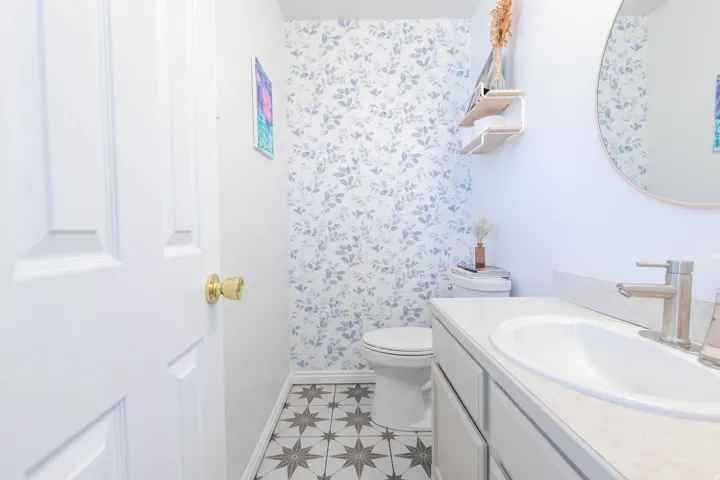 Powder room featuring patterned floor tile, a single vanity with a white countertop and integrated sink, a large round mirror, and a wallpaper accent wall