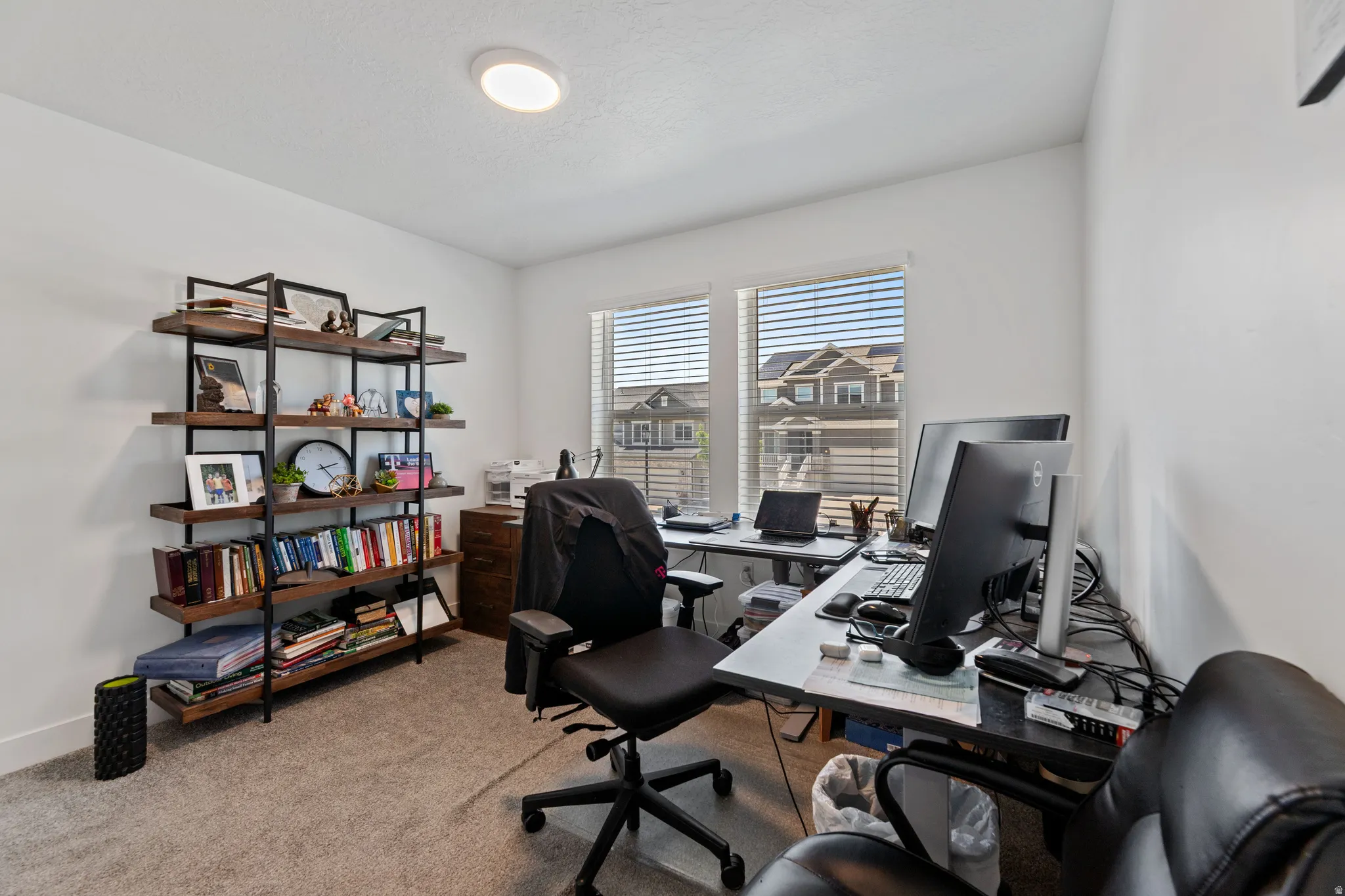 Dedicated workspace featuring two large windows, recessed ceiling lighting, wall-to-wall carpeting, and a built-in shelving unit with dark wood-finish shelves and a black metal frame