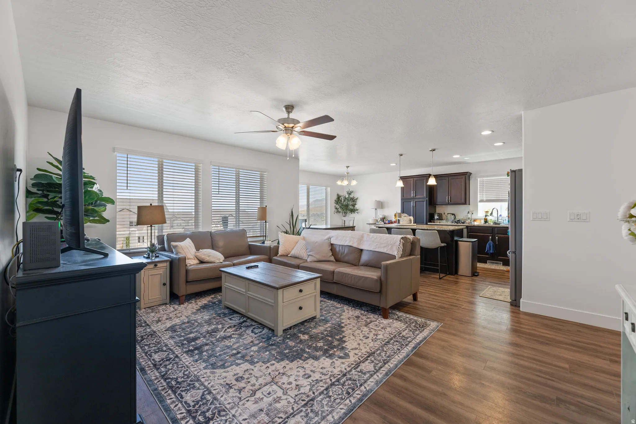 Open-concept living space featuring wood-finish flooring throughout, a ceiling fan with integrated lighting, and multiple windows with horizontal blinds
