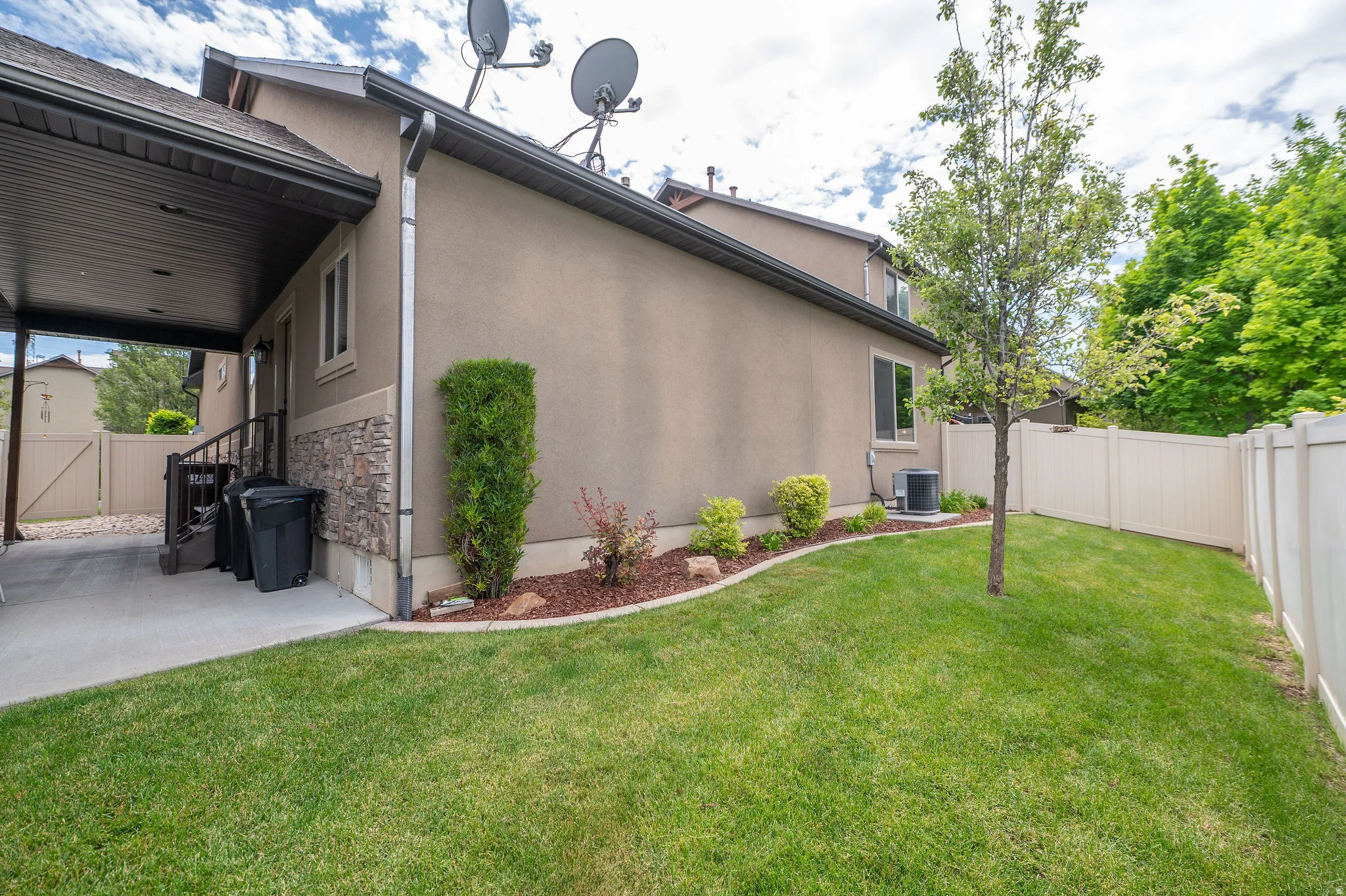 Back of the home shows stucco and stacked stone exterior, covered patio, landscaped yard, and vinyl privacy fence. Low-maintenance and well-kept.
