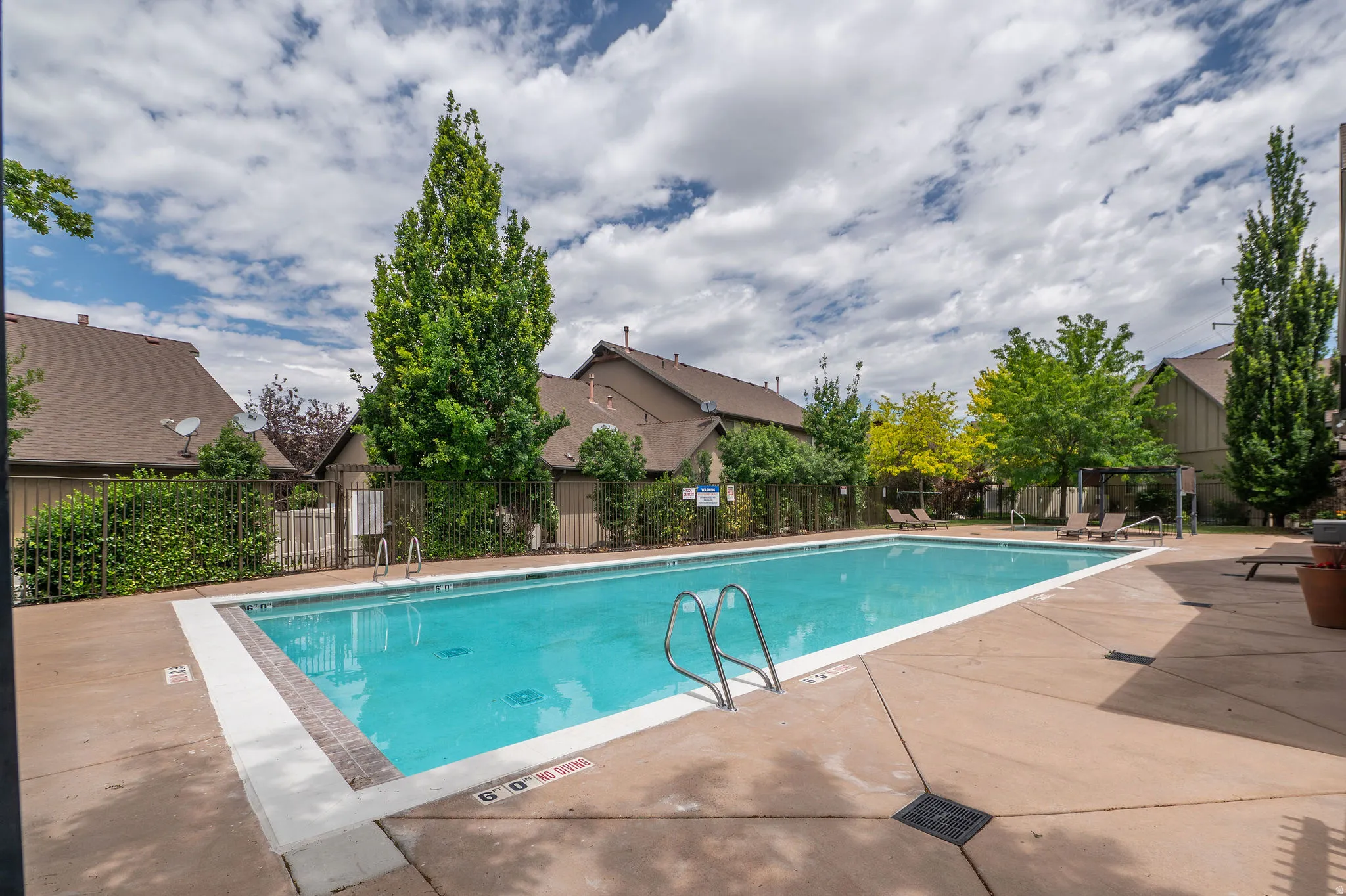 Sparkling community pool at ground level, surrounded by mature trees, iron fencing, lounge chairs, and a pergola seating area. Blue skies and clean water make this the place to be all summer.