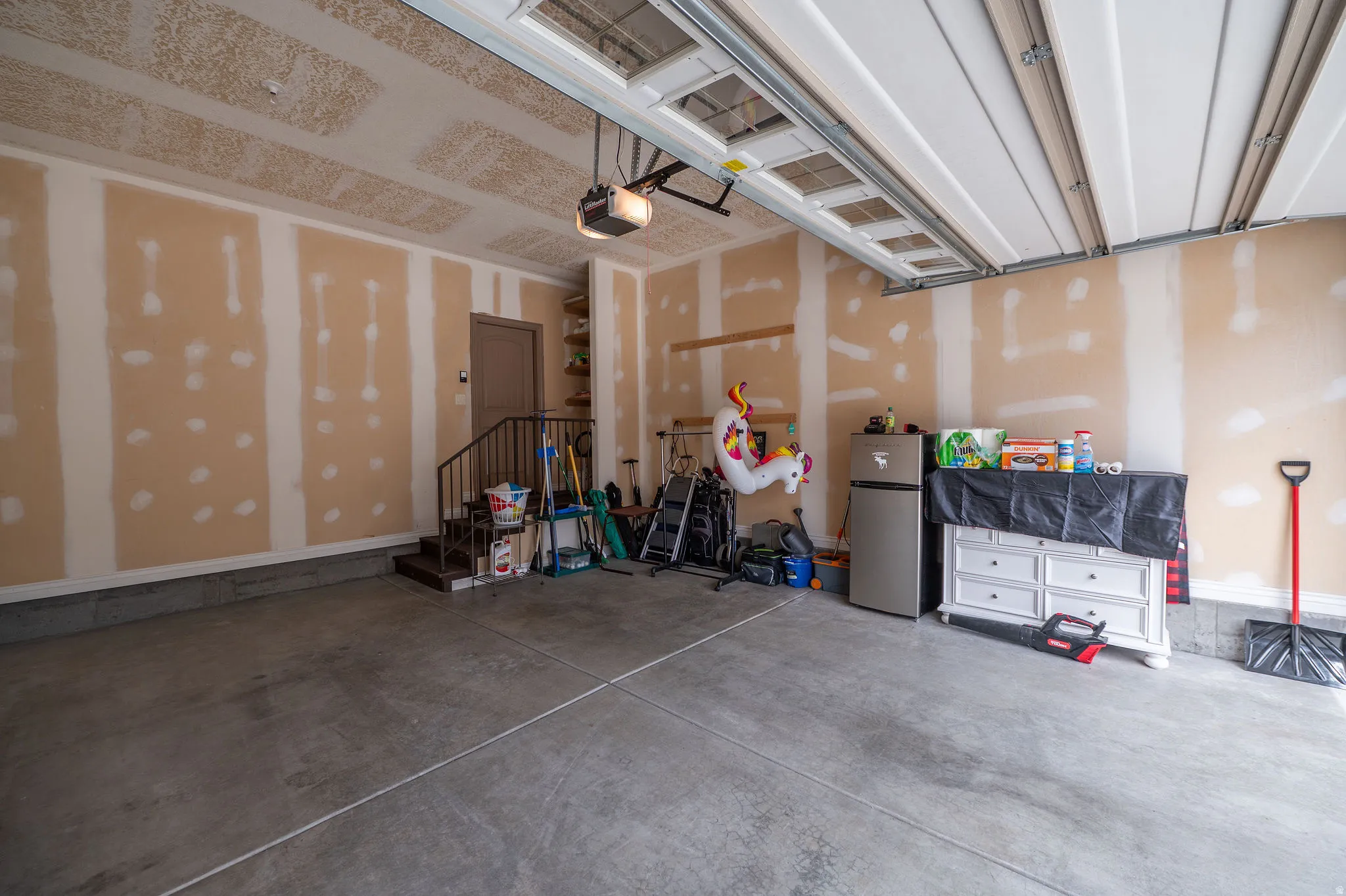 Second look at the spacious two-car garage showing the full depth, drywalled walls, automatic opener, built-in shelving, and direct interior access via staircase entry.