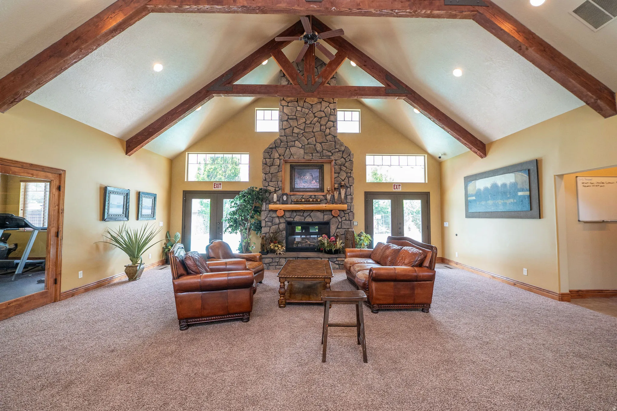 Stunning clubhouse great room with soaring vaulted ceilings, dramatic exposed wood beam trusses, floor-to-ceiling stacked stone fireplace, and French doors opening to the pool area. The fitness room is visible off to the side. This amenity space is except