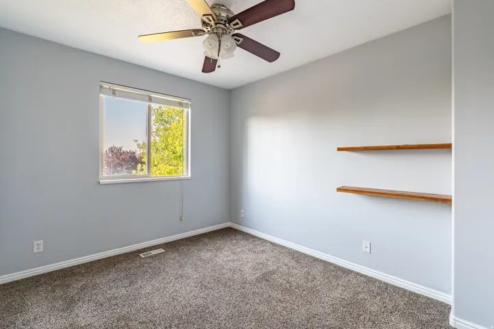 Room featuring a window with exterior tree scenery, light gray walls, textured carpet flooring, and a ceiling fan with integrated lighting