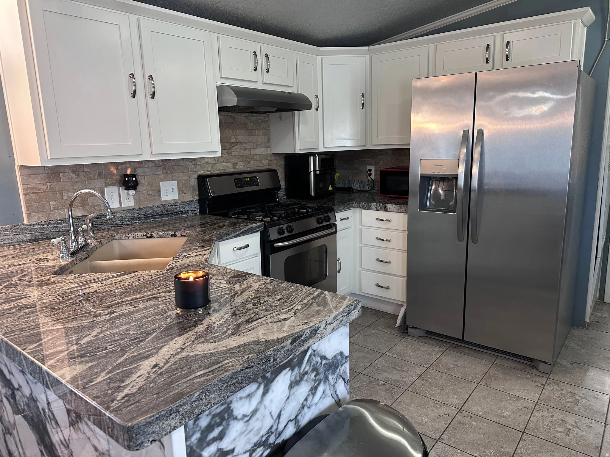 Updated kitchen featuring white shaker cabinetry, patterned stone countertops, stainless steel appliances, a tile backsplash, and tile flooring