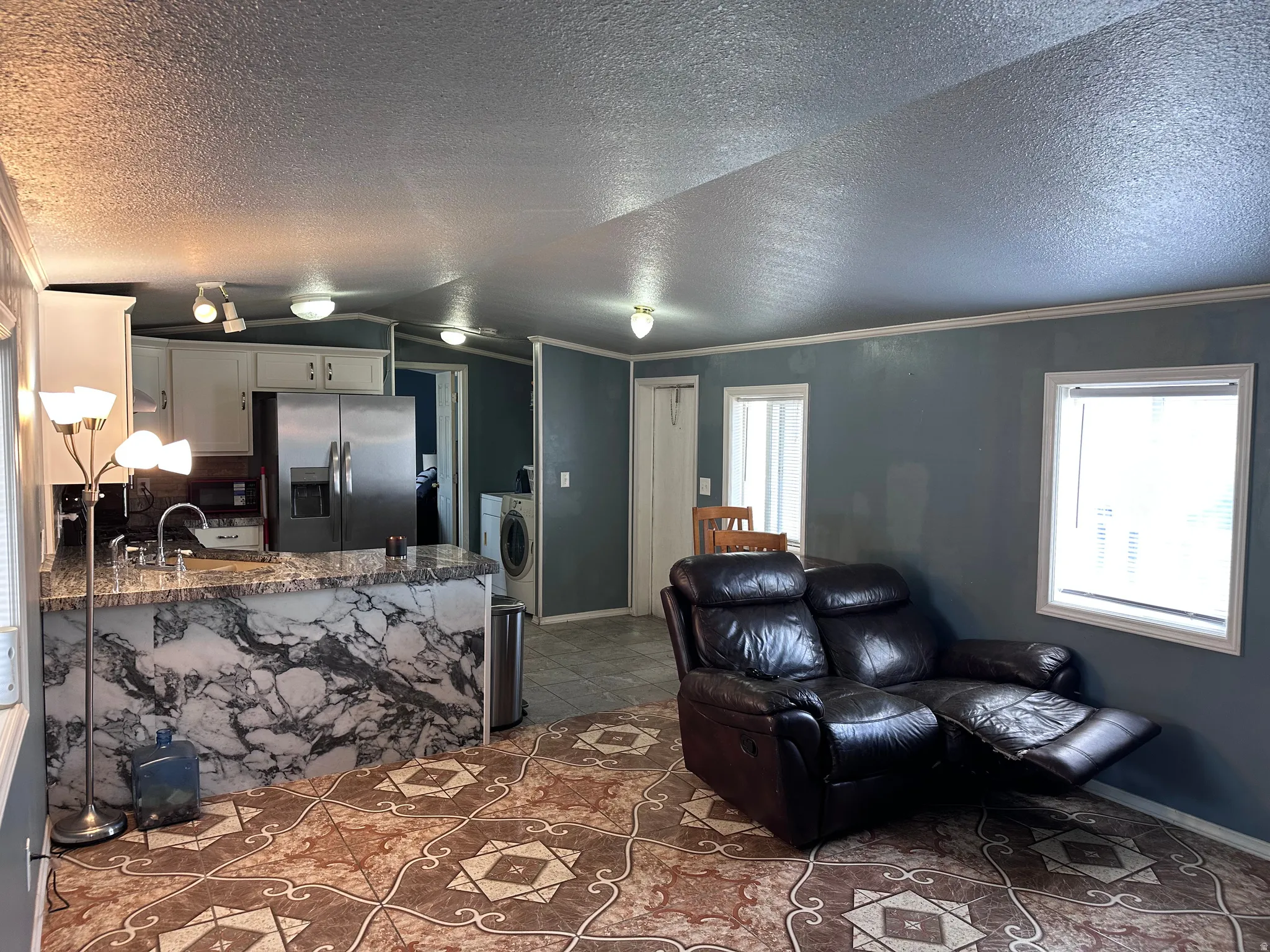 Kitchen and living area featuring patterned tile flooring, an eat-at kitchen island with a marbled finish, white cabinetry, and stainless steel appliances