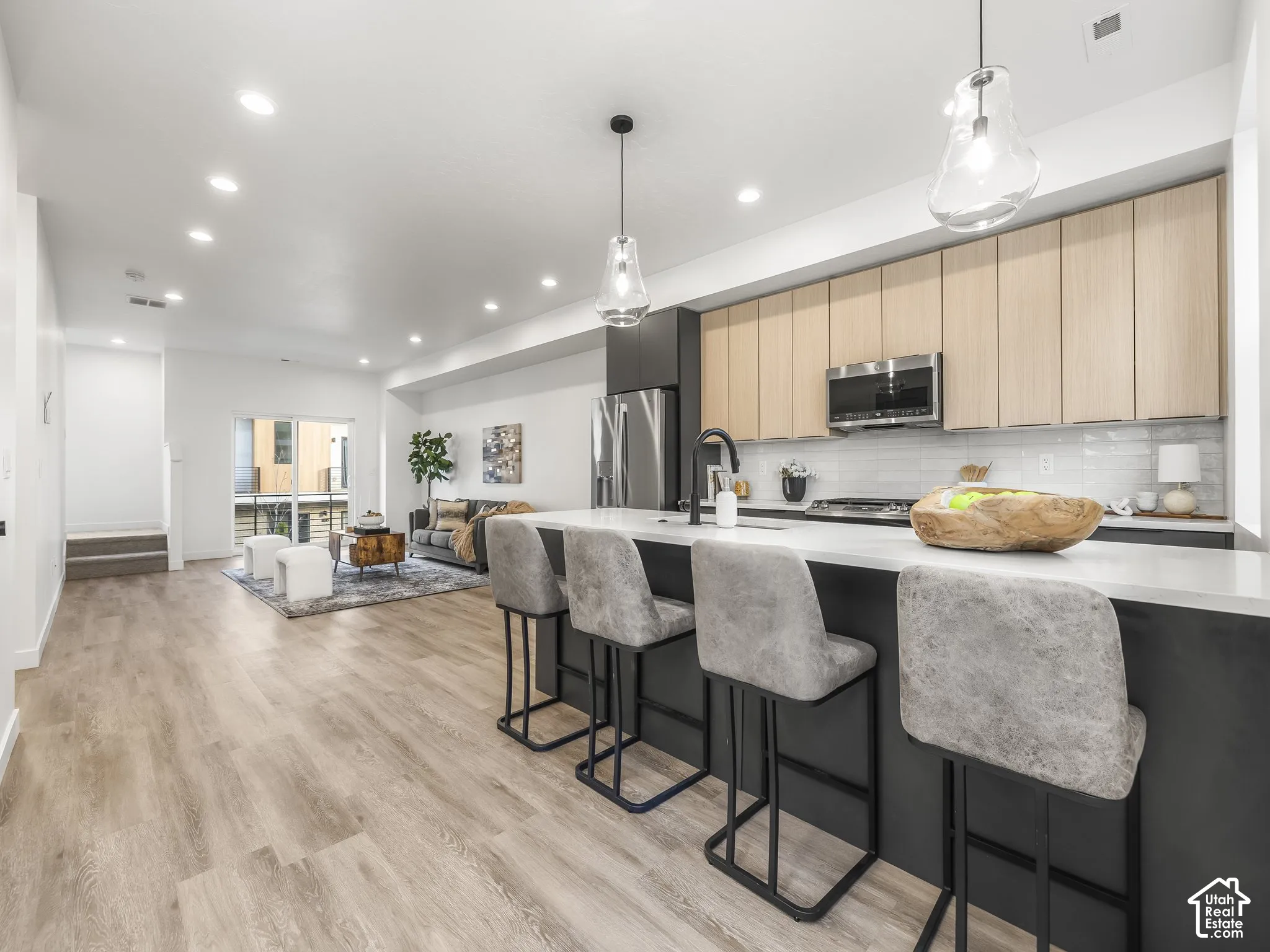 Open-concept living space featuring a contemporary kitchen with light wood-finish cabinetry, a large center island with a white countertop, and stainless steel appliances