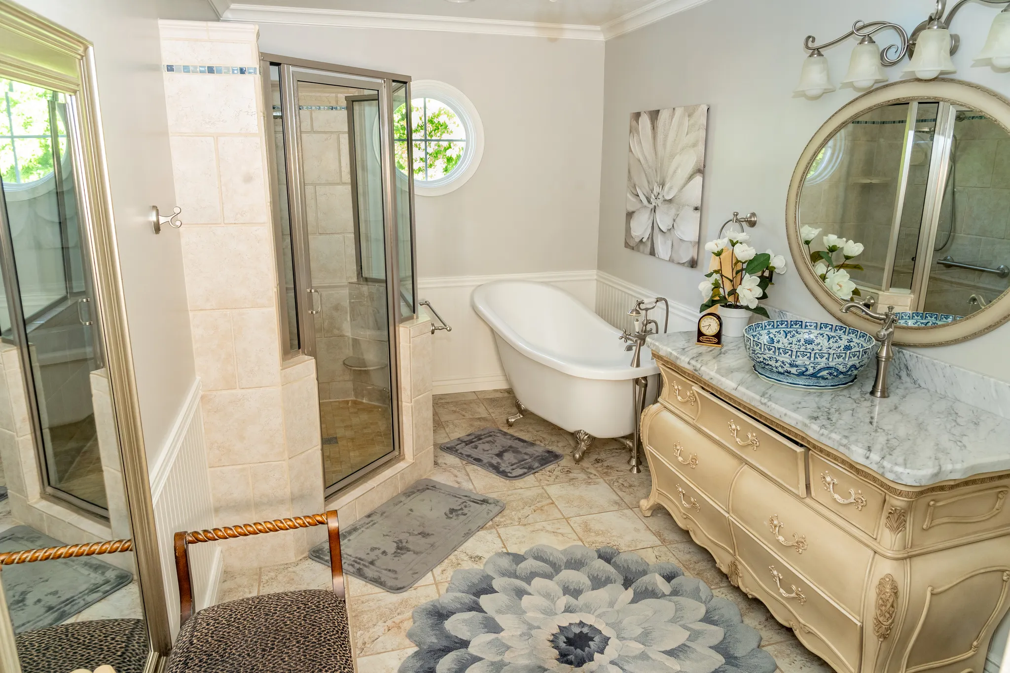 Bathroom featuring a walk-in shower with glass enclosure, a freestanding clawfoot tub, a vanity with a stone countertop and decorative vessel sink, a circular mirror, and a round window