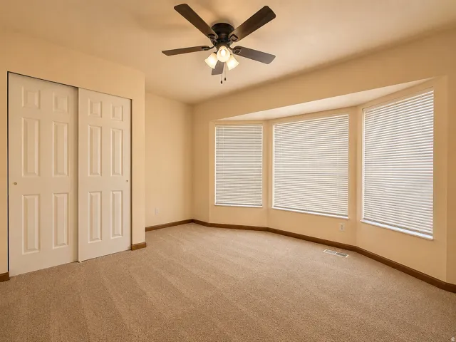 Carpeted room featuring a ceiling fan with integrated lighting, a bay window with three vertical blinds, and a built-in closet with bi-fold doors